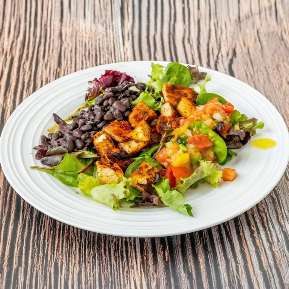 A vibrant salad with grilled chicken, black beans, mixed greens, and pico de gallo on a white plate.